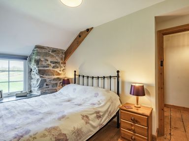 A bedroom with a bed, a wooden nightstand with a lamp, stone wall section, window, and wooden floor at Ty Capel Jeriwsalem Bronaber near Trawsfynydd