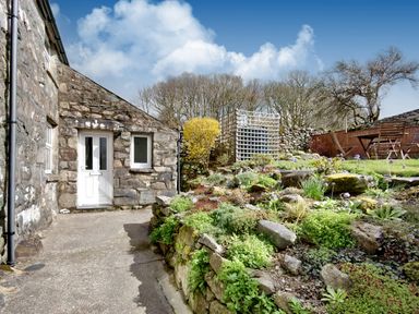 Stone house exterior with white door and window a concrete path and garden with plants and wooden table and chairs at Ty Capel Jeriwsalem in Bronaber near Trawsfynydd