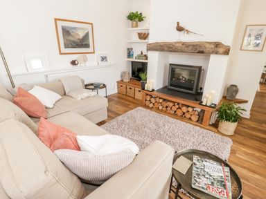 A living room with a beige sofa a fireplace with firewood and a rug at Beudy Hywel in Llanrug near Llanberis