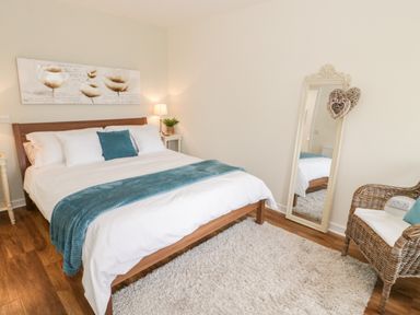 A bedroom with a wooden bed with white and teal bedding a white rug a full length mirror and a wicker chair with a cushion at Beudy Hywel in Llanrug near Llanberis