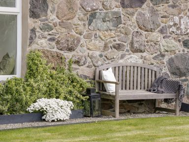 A wooden bench with a white pillow and a black and white blanket next to a stone wall and greenery at Beudy Hywel in Llanrug near Llanberis