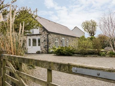 A stone house with white doors and windows surrounded by bushes and trees behind a wooden gate at Beudy Hywel in Llanrug near Llanberis