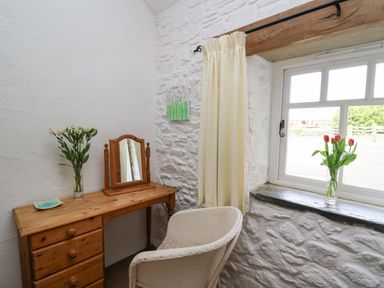 A small corner with a wooden dressing table and mirror a white chair two vases with flowers and a window with a curtain at Bwthyn Bach in Newport