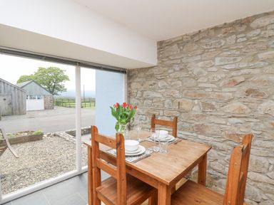 A dining area with a wooden table and chairs set for two next to a stone wall and large window showing an outdoor scene at Y Cwtch in Newport