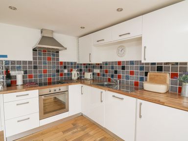 A kitchen with wooden floors white cabinets a stainless steel oven and hood tiled backsplash with red gray and blue tiles a sink kettle toaster and a bread box at Y Cwtch in Newport