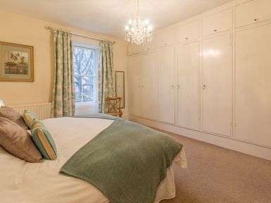 A bedroom with a bed covered in pillows and a blanket a window with curtains a chair a wall mirror and built in wardrobes at The Old Exchange Corbridge near Hexham