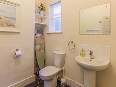 A bathroom with a toilet, sink with soap, mirror, window, ironing board, and painting at The Old Exchange in Corbridge near Hexham