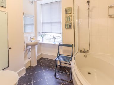 A bathroom with a bathtub shower combination a sink toilet a window with blinds a black chair and decorative items at The Old Exchange in Corbridge near Hexham