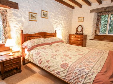 A bedroom with a wooden bed and floral bedspread beside windows and wooden furniture at Nant Cottage in Llanrwst near Betws-y-Coed