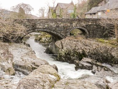 A stone arch bridge over a river with rocks and buildings in the background at Nant Cottage in Llanrwst near Betws-y-Coed