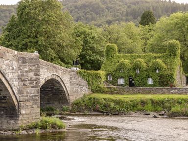 A stone bridge over a river with a house covered in green ivy next to trees and hills at Nant Cottage in Llanrwst near Betws-y-Coed