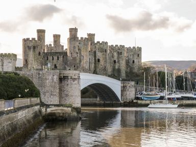 A stone castle with multiple towers next to a bridge over a body of water with sailboats docked nearby