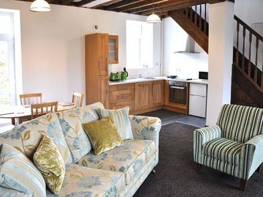 A living room with a floral sofa striped armchair wooden dining table and an open kitchen under wooden stairs at Hendre Aled Cottage 3 in Llansannan
