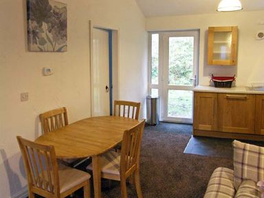 A dining area with a wooden table and four chairs next to a kitchen with wooden cabinets and glass door at Hendre Aled Cottage 1 in Llansannan near Llanrwst