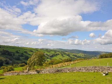 A rural landscape with green hills trees and a stone wall under a partly cloudy sky at Hendre Aled Cottage 1 Llansannan near Llanrwst