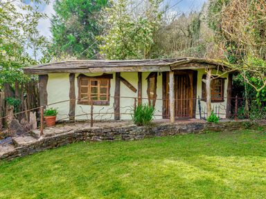 A small rustic house with wooden beams windows and a door surrounded by greenery and a grassy lawn at Brookbank Folly in Mitcheldean