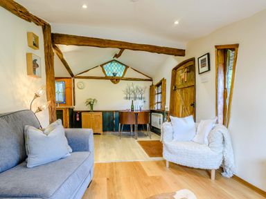 A room with a gray sofa and a white armchair wooden beams a wooden door a dining table with chairs and cabinets at Brookbank Folly in Mitcheldean