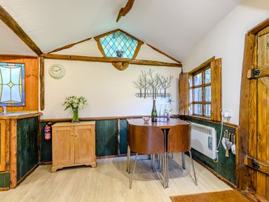 A small dining area with a wooden table and four chairs next to a window and a wooden cabinet with flowers at Brookbank Folly in Mitcheldean