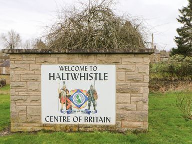 A stone wall with a welcome sign to Haltwhistle Centre of Britain surrounded by grass and trees