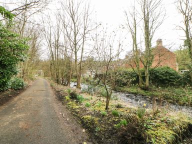 A pathway next to a river with leafless trees and brick houses in the background at Hollie Cottage in Haltwhistle