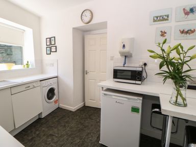 A kitchen with a washing machine dishwasher small fridge microwave flowers and a clock on the wall at Hollie Cottage in Haltwhistle