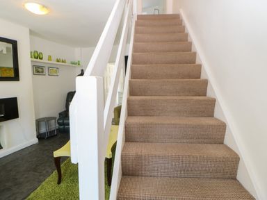 A staircase with carpet leading to an upper floor with a partial view of a living room with a yellow table green rug and a black chair at Hollie Cottage in Haltwhistle