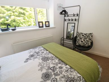 A bedroom with a bed covered in floral sheets and a green throw a window with decorative ducks two framed pictures a black metal clothes rack with a mirror and hangers and a black chair with a patterned cushion at Hollie Cottage in Haltwhistle