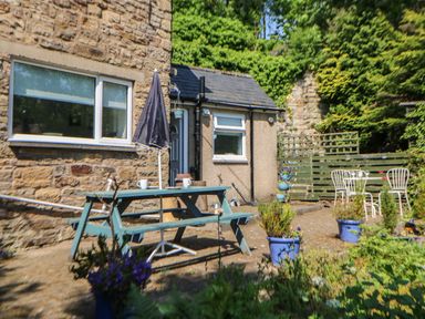 An outdoor patio area with a wooden picnic table with umbrella two white metal chairs potted plants and a stone building at Hollie Cottage in Haltwhistle