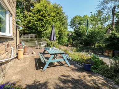 An outdoor patio with a picnic table and umbrella next to a garden and stone house at Hollie Cottage in Haltwhistle