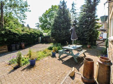 An outdoor patio area with a wooden picnic table and umbrella surrounded by potted plants and trees at Hollie Cottage in Haltwhistle