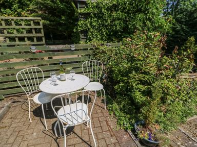 A white metal table and three chairs on a paved patio with a bottle of wine and four glasses surrounded by green plants and a wooden fence at Hollie Cottage in Haltwhistle