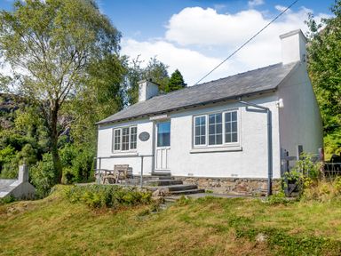 A white cottage with two chimneys and steps leading to the door surrounded by trees at Penlan in Beddgelert
