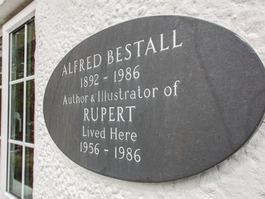 A black plaque on a white wall next to a window commemorating Alfred Bestall author and illustrator of Rupert at Penlan in Beddgelert