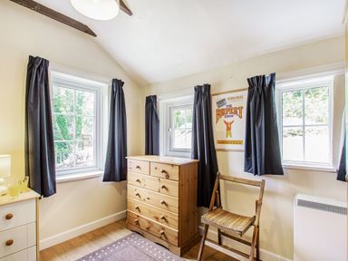 A small room with three windows with dark curtains a wooden chest of drawers a wooden chair a small cabinet and a framed poster on the wall at Penlan in Beddgelert