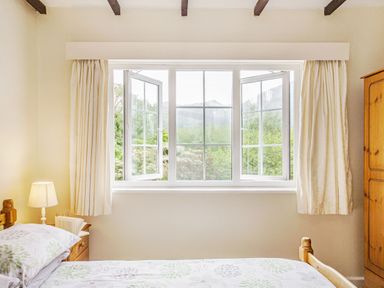 A bedroom with a wooden bed a bedside table with a lamp and book a wooden wardrobe and an open window with cream curtains at Penlan in Beddgelert