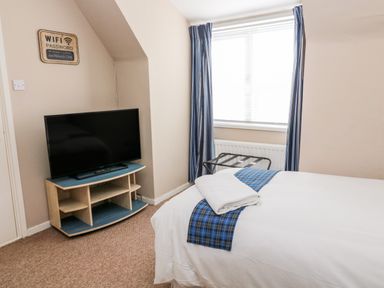 A bedroom with a bed having a plaid runner and towel near a window with blue striped curtains and a television on a corner stand at Cra-na-ge in Embleton