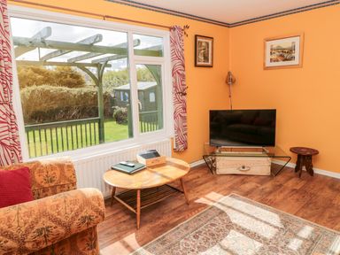 A living room with a wooden coffee table a patterned armchair a flat screen tv on a glass stand yellow walls and a large window showing a garden at Cra-na-ge in Embleton