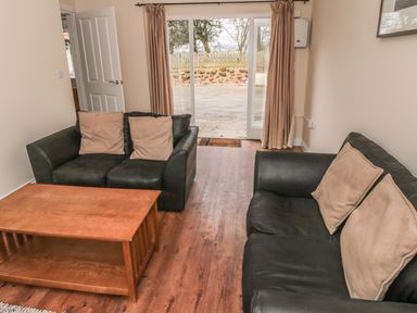 A living room with two black sofas with beige cushions a wooden coffee table wooden floor and glass sliding doors with curtains at No 1 Low Hall Cottages Scalby near Scarborough