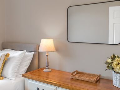 A bedside table with a lamp a wicker tray and a flower pot next to a bed with pillows and a beige headboard at Colt Park Cottage in Ulverston