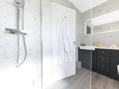 A bathroom with a shower area a white door with a white robe hanging a black cabinet with a sink and mirror at Colt Park Cottage in Ulverston