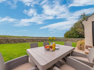An outdoor patio with a table and chairs set with fruit juice and flowers near a stone wall at Colt Park Cottage in Ulverston
