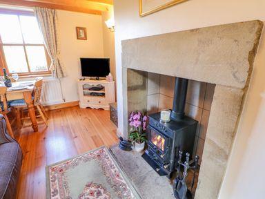 A room with a wood-burning stove in a stone fireplace a TV on a white stand a wooden dining table and chairs and a floral rug on wooden floor Moorgair Cottage Slaley near Hexham