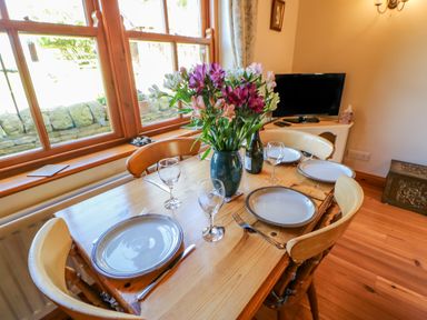 A dining table set with plates, glasses, cutlery, and a vase of flowers near a window at Moorgair Cottage in Slaley near Hexham