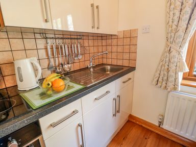 A kitchen countertop with a sink a kettle a bowl of fruit cooking utensils hanging and a window with floral curtains at Moorgair Cottage in Slaley near Hexham
