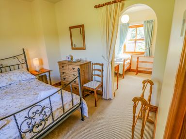 A bedroom with a metal bed frame a wooden dresser a chair and a curtained archway leading to a smaller room with a window and another wooden bed at Moorgair Cottage in Slaley near Hexham