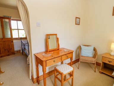 A bedroom with a wooden dressing table and stool a wicker chair with a cushion and a bedside table with a lamp at Moorgair Cottage in Slaley near Hexham