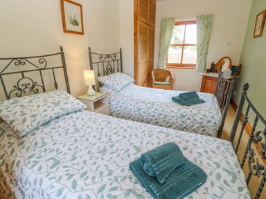 A bedroom with two single beds with patterned bedding a nightstand with a lamp a wicker chair a window with curtains and a wooden dresser with a mirror at Moorgair Cottage in Slaley near Hexham