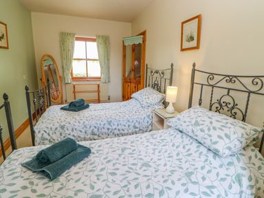 A bedroom with two single beds with patterned bedding a bedside table with lamp a wooden door a window with curtains and a floor mirror at Moorgair Cottage in Slaley near Hexham