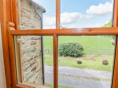 A wooden window looking out to a stone wall, garden with bushes, flower pots, and a green field at Moorgair Cottage in Slaley near Hexham