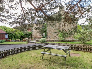 A garden with a wooden picnic table surrounded by a low wooden fence and a stone cottage in the background at Moorgair Cottage Slaley near Hexham